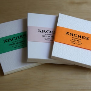 May include: Three stacks of white Arches watercolor paper, each wrapped in a colored paper band. The green band reads Cold Pressed, the light pink band reads Hot Pressed, and the orange band reads Rough. Each pack specifies 300 g/m² and 100% cotton.