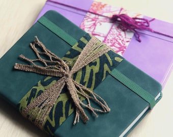 May include: A dark teal notebook with a green elastic band and a decorative band featuring a brown and green abstract pattern tied with brown twine. Behind it lies a light purple notebook with a pink and white patterned band.