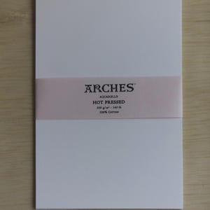 May include: A rectangular pad of white Arches Aquarelle hot pressed watercolor paper featuring a light pink paper band across the center. The band displays the text Arches, Aquarelle, Hot Pressed, 300 g/m&sup2; - 140 lb, and 100% Cotton in black.