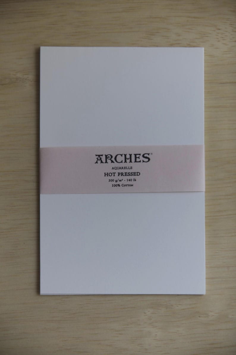 May include: A rectangular pad of white Arches Aquarelle hot pressed watercolor paper featuring a light pink paper band across the center. The band displays the text Arches, Aquarelle, Hot Pressed, 300 g/m&sup2; - 140 lb, and 100% Cotton in black.