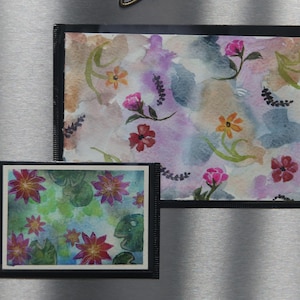 May include: Two rectangular magnetic frames with black borders. The larger frame displays a watercolor painting of pink, orange, and red flowers on a soft purple and blue wash. The smaller frame shows a watercolor of pink water lilies on green pads.
