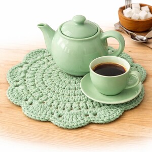 May include: A light green tea set comprising a teapot, teacup, and saucer, resting on a crocheted doily. The teacup contains a dark liquid, with a small wooden bowl of sugar cubes in the background. The set is on a wooden surface.