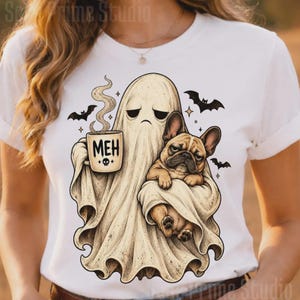 Könnte beinhalten: Ein weißes T-Shirt mit einer grafischen Illustration eines traurigen Geistes, der eine Kaffeetasse mit dem Text MEH und einem kleinen Totenkopf-Symbol hält. Der Geist hält eine braune französische Bulldogge. Mehrere kleine schwarze Fledermäuse und Sterne umgeben die Figuren.