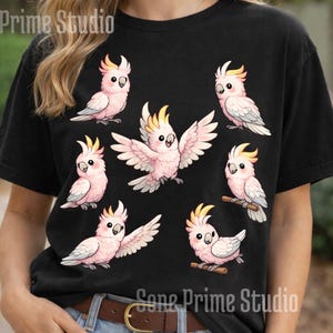 May include: Black t-shirt featuring a design of several cartoon cockatoos in various poses. The birds are primarily pink with yellow crests and gray wings. The text "Prime Studio" is printed at the top and bottom of the shirt.