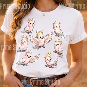 May include: White t-shirt featuring a design of cartoon cockatoos in various poses. The birds are primarily pink and white with yellow crests and gray wings. The design includes several cockatoos, some perched and others with wings spread.
