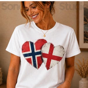 Può includere: Una t-shirt bianca a maniche corte con due grafiche a forma di cuore sovrapposte sul petto. Il cuore a sinistra mostra la bandiera blu, bianca e rossa della Norvegia, mentre il cuore a destra mostra la bandiera rossa e bianca dell'Inghilterra.