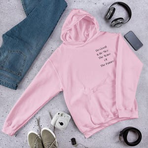 May include: A pale pink hoodie with the text "Do Good & Be Nice The Wave Of The Future" printed on the front. The hoodie has a kangaroo pocket and a hood. Blue jeans, trainers, headphones, and a camera are also in the image.
