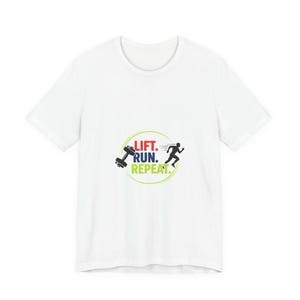 Puede incluir: Camiseta blanca con un diseño gráfico de temática fitness. El diseño incluye una pesa, una figura corriendo y las palabras "LIFT. RUN. REPEAT." en rojo, verde y negro. La camiseta está hecha de una tela suave y clara.