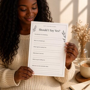 May include: A white paper with the text "Should I Say Yes?" and prompts for decision-making. The paper is held by a person wearing a cream-colored sweater. The paper has a floral design and the words "Next Chapter Studio".