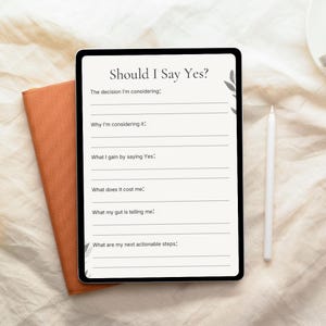 May include: A digital tablet displaying the text "Should I Say Yes?" with prompts for decision-making. The tablet is next to an orange notebook and a white stylus. The background is a light-colored fabric.