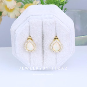 May include: A pair of gold-colored teardrop earrings with a white opal center stone, surrounded by a halo of small, clear crystals. The earrings are displayed on a white, textured jewelry box. The text "JEWELRYBYITEZAZ" is visible.