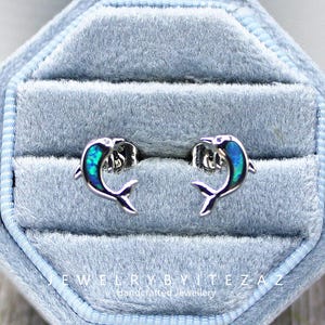 May include: Silver dolphin stud earrings with blue and green iridescent inlay, presented in a light blue velvet jewellery box. The text "JEWELRYBYITEZAZ Handcrafted Jewellery" is visible.