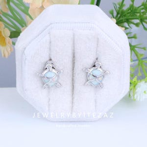 Könnte beinhalten: Silberfarbene Schildkröten-Ohrstecker mit hellblauem, opalartigem Mittelteil, präsentiert in einer weißen Schmuckschatulle. Die Ohrringe haben detaillierte Flossen- und Kopfdesigns. Der Text "JEWELRYBYTEZAZ" und "Handcrafted Jeweller" sind sichtbar.
