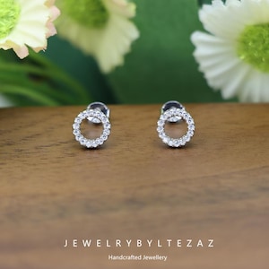 May include: A pair of silver stud earrings with a circular design. Each earring features a ring of small, clear gemstones. The earrings are displayed on a wooden surface, with the text "JEWELRYBYLTEZAZ" and "Handcrafted Jewellery" below.