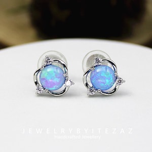 May include: A pair of silver stud earrings with a round, iridescent blue opal center stone. Each earring features a delicate silver design with three small, clear accent stones. The earrings are displayed on a white surface.