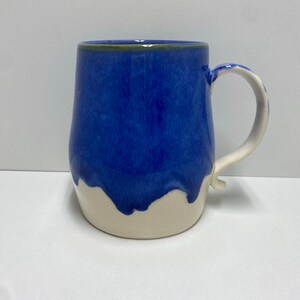 May include: A handmade ceramic mug with a blue glaze that drips down over a cream-colored base. The mug has a curved handle and a smooth, glossy finish. The mug is designed for hot beverages.