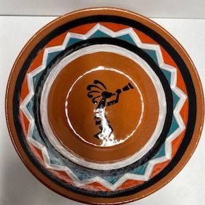 May include: A round, ceramic plate with a Kokopelli design. The plate features concentric rings of color, including orange, black, white, and turquoise. The Kokopelli figure is black and is playing a flute. The plate has a glossy finish.