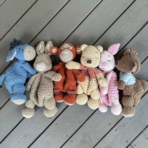 May include: A collection of seven handmade crochet stuffed animals, including a blue donkey, a tan bunny, an orange tiger, a yellow bear, a pink pig, and a brown bear. The toys are arranged on a gray wooden surface.