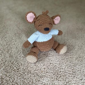 May include: A brown crocheted stuffed animal shaped like a bear with a light blue shirt, pink inner ears, and beige feet. The bear has a dark brown nose and closed eyes, with a small tuft of brown yarn on top of its head, crafted in a soft texture.