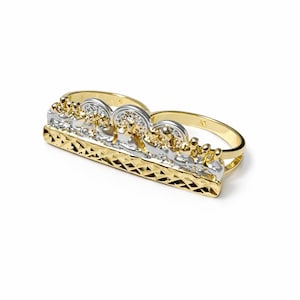 May include: A two-finger ring with a gold band and a detailed centerpiece. The centerpiece features a gold and silver design with intricate detailing. The ring is set against a white background.
