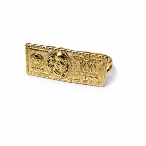 May include: A gold-colored ring featuring a detailed design of a $100 bill. The ring showcases the portrait of Benjamin Franklin and the number 100. The ring is designed to be worn across two fingers.