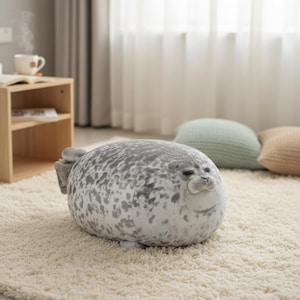 Può includere: Un cuscino a forma di foca grigia e bianca è appoggiato su un tappeto color crema. La foca ha un viso sorridente e pinne. Lo sfondo comprende un tavolino di legno, una finestra e cuscini decorativi.
