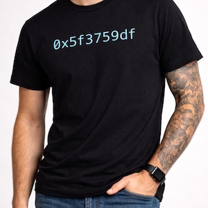 Puede incluir: Camiseta negra de manga corta con el texto azul claro "0x5f3759df". La persona que lleva la camiseta tiene un tatuaje en el brazo y un reloj negro. También lleva vaqueros azules.