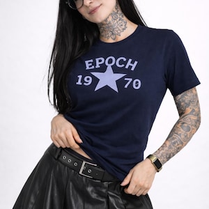 Può includere: T-shirt blu navy con la scritta "EPOCH 1970" e una stella. L'outfit comprende una gonna a pieghe nera e una cintura nera. La persona indossa occhiali e un orologio.