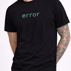 Può includere: T-shirt nera con la scritta "error" in verde, centrata sul petto. La maglietta a maniche corte è abbinata a jeans blu. La persona indossa un tatuaggio sul braccio.