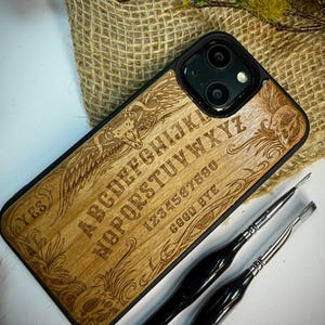 May include: A wooden phone case with a black border, featuring an engraved Ouija board design. The design includes letters, numbers, and the words "YES" and "GOOD BYE". Two black-handled paint brushes are in the foreground.