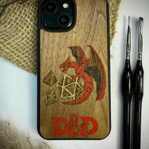 May include: Wooden phone case with a black border, featuring a red dragon, dice, and the letters "D&D". The case has a natural wood grain finish. Two black-handled paint brushes are on the right side of the image.