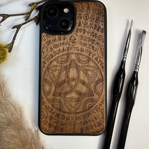 May include: A wooden phone case with a black border, featuring an engraved Celtic knot design and surrounding symbols. Two black-handled tools are positioned to the right of the case. The case is made of light brown wood.