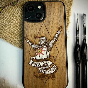 May include: Wooden iPhone case with a black border. The case features a detailed engraving of a knight with arms raised, a sun emblem, and the text "Praise the Sun" on a banner. Three black paint brushes are on the right.