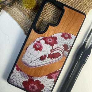 Può includere: Una custodia per telefono con retro in legno. Presenta un'illustrazione dettagliata di un serpente con squame bianche e motivi floreali rossi. La custodia ha un bordo nero e uno sfondo con venature del legno naturale.