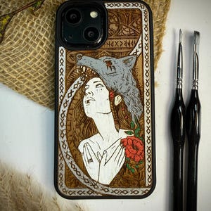 Könnte beinhalten: Schwarze Handyhülle mit detailliertem Holzbranddesign. Das Kunstwerk zeigt eine Frau mit Wolfskopfschmuck, eine rote Rose und komplizierte Muster. Zwei Pinsel sind auf der rechten Seite des Bildes zu sehen.