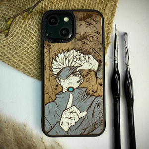 Puede incluir: Funda de teléfono negra con diseño de vetas de madera que presenta una ilustración de un personaje de anime. El personaje está representado en tonos grises y azules, con una mano levantada hacia la cara. Dos pinceles negros están al lado de la funda.