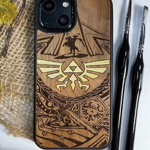 May include: Wooden phone case with a black border, featuring a laser-etched design of a video game theme. The design includes a silhouette of a figure, a crest, and other graphic elements. Two black paint brushes are next to the phone case.