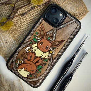 May include: A wooden phone case featuring a carved Eevee design with white flowers and a black border. The case is brown with a black camera lens area. Two black paint brushes are next to the phone case.