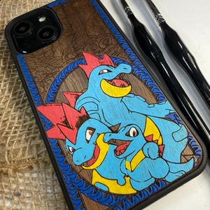 May include: A wooden phone case featuring a blue, red, and yellow cartoon design. The case has a black border and a detailed wood grain pattern. The design includes three cartoon characters with red crests and yellow accents.