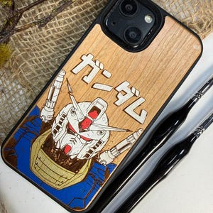 Könnte beinhalten: Handyhülle aus Holz mit einer Roboterillustration und japanischem Text. Die Hülle hat eine natürliche Holzmaserung und einen schwarzen Rand. Das Roboterdesign ist in Blau, Gold und Rot gehalten. Zwei schwarze Pinsel sind sichtbar. Der japanische Text lautet "ガンダム".