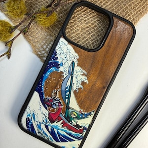 Op de afbeelding: Telefoonhoesje met een houten achterkant en zwarte rand. De hoes heeft een kleurrijke illustratie van een boot die op een golf vaart, met een draak en een personage dat een zeil vasthoudt. Het ontwerp is geïnspireerd op Japanse kunst.