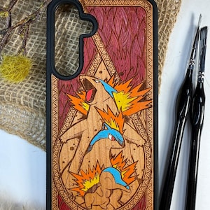 Puede incluir: Una funda de teléfono con una parte trasera de madera con un diseño grabado con láser de tres personajes de dibujos animados con llamas. La funda tiene un borde negro y un fondo rojo y marrón. El diseño está rodeado por un borde decorativo.