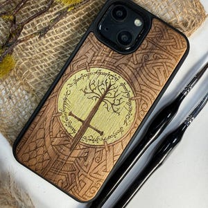 May include: A wooden phone case with a detailed tree and sword design, featuring engraved text around a central emblem. The case has a black border and a natural wood grain finish. Two black paint brushes are visible.