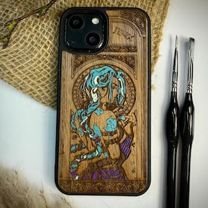 K&ouml;nnte beinhalten: Holz-Handyh&uuml;lle mit schwarzem Rand und detailliertem Laser-Gravur-Design. Das Design zeigt eine Figur mit blauen Haaren und dem Wort "JINX" oben. Zwei schwarze Pinsel liegen daneben.