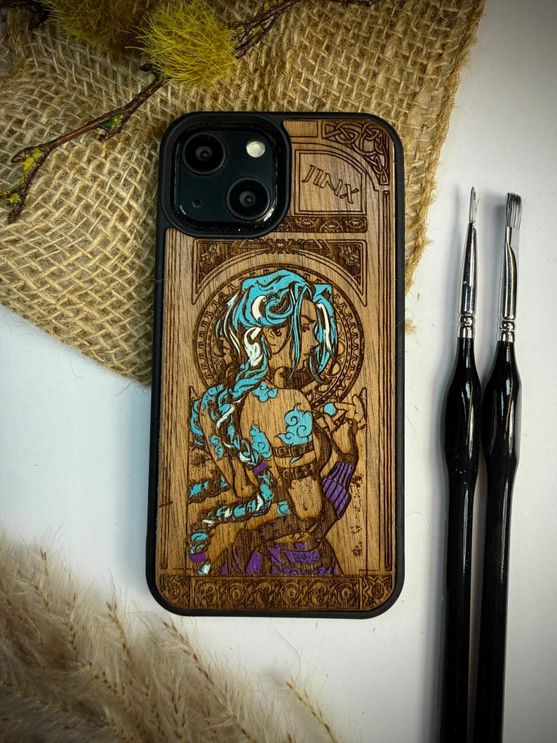 K&ouml;nnte beinhalten: Holz-Handyh&uuml;lle mit schwarzem Rand und detailliertem Laser-Gravur-Design. Das Design zeigt eine Figur mit blauen Haaren und dem Wort "JINX" oben. Zwei schwarze Pinsel liegen daneben.
