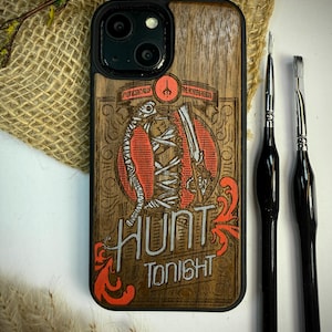 May include: Wooden phone case with a carved design featuring a skeleton, a gun, and the words "HUNT TONIGHT" in white and orange. The case has a black border and is next to two black paint brushes.