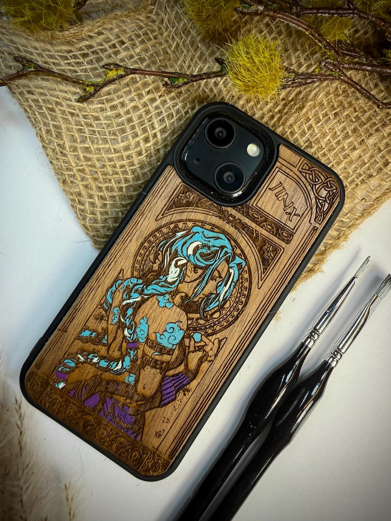 K&ouml;nnte beinhalten: Handy-H&uuml;lle aus Holz mit einem lasergravierten Design eines Charakters mit blauen Haaren und Akzenten. Die H&uuml;lle hat einen schwarzen Rand und der Name "Jinx" ist sichtbar. Das Kunstwerk ist in Braun-, Blau- und Lilat&ouml;nen gehalten.