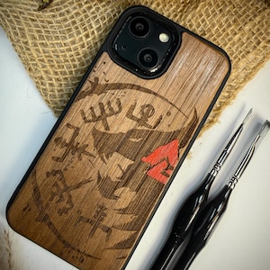 Puede incluir: Funda de teléfono de madera con borde negro, con un diseño tallado y un acento rojo. La funda se muestra con dos pinceles negros, sobre un fondo de arpillera.