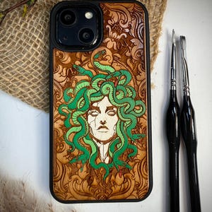 May include: A black phone case with a Medusa design. The case has a detailed wood-burned image of Medusa with green snakes and a carved floral pattern. Two black paint brushes are next to the case.