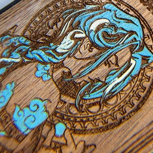 K&ouml;nnte beinhalten: Nahaufnahme einer Holzoberfl&auml;che mit detailliertem Laser-&Auml;tz-Design. Das Design zeigt eine blau-wei&szlig;e Wasserkreatur in einem kreisf&ouml;rmigen Rahmen. Das Holz hat einen nat&uuml;rlichen Braunton, und das Design ist aufwendig geschnitzt.